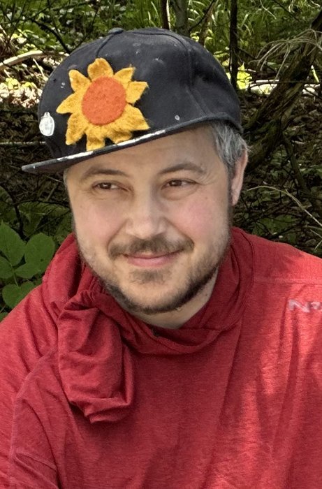 Portrait of Encian Pastel, smiling, wearing a black cap with a sunflower patch and a red hoodie, outdoors surrounded by greenery
